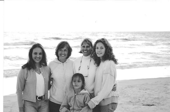 Five women and a child smile together on a beach with waves gently lapping at the shore.