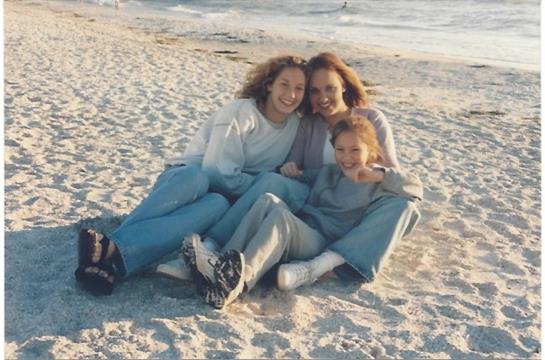 Three young women sit together on the sandy beach, sharing laughter and joy as the sun sets.
