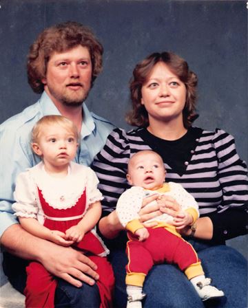 A family of four poses together, two adults holding two young children, in a studio.