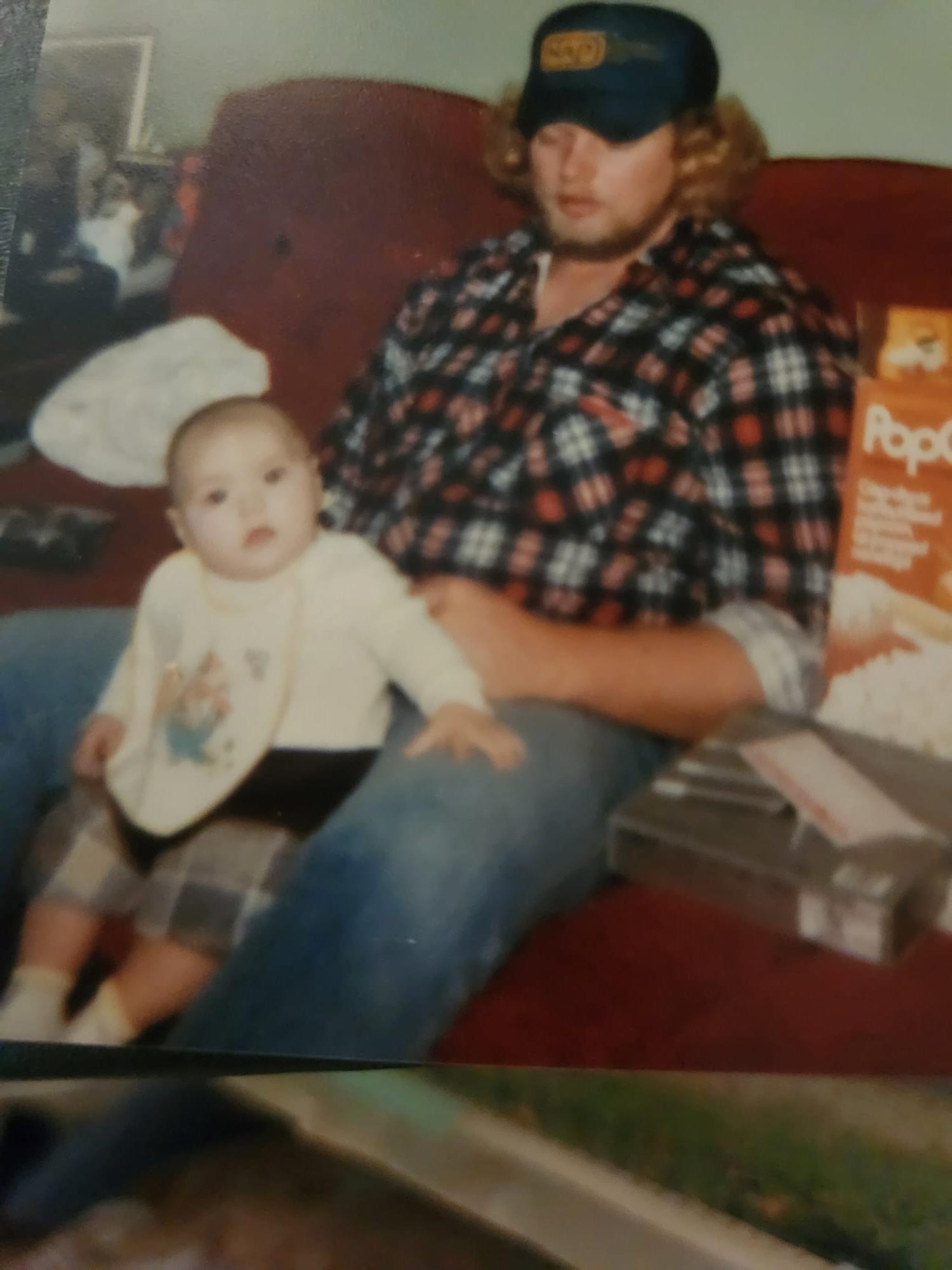 A man in plaid shirt holds a baby on his lap in a relaxed living room filled with snacks.