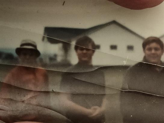 Three friends stand close together in an outdoor setting, with a barn in the background.