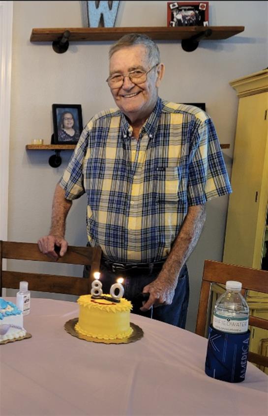 A joyful man stands beside a yellow birthday cake with candles, celebrating his 80th birthday.