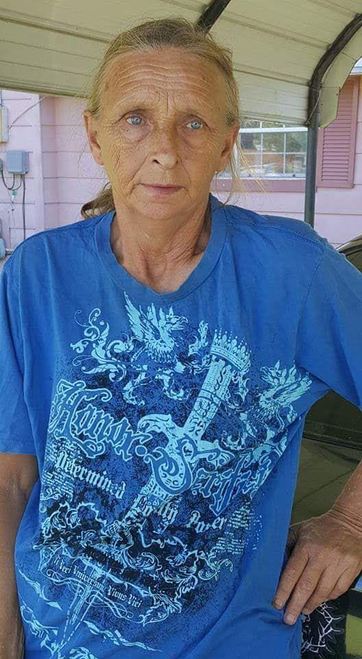 Individual with blue eyes and long hair poses by a car outside a house on a sunny day.
