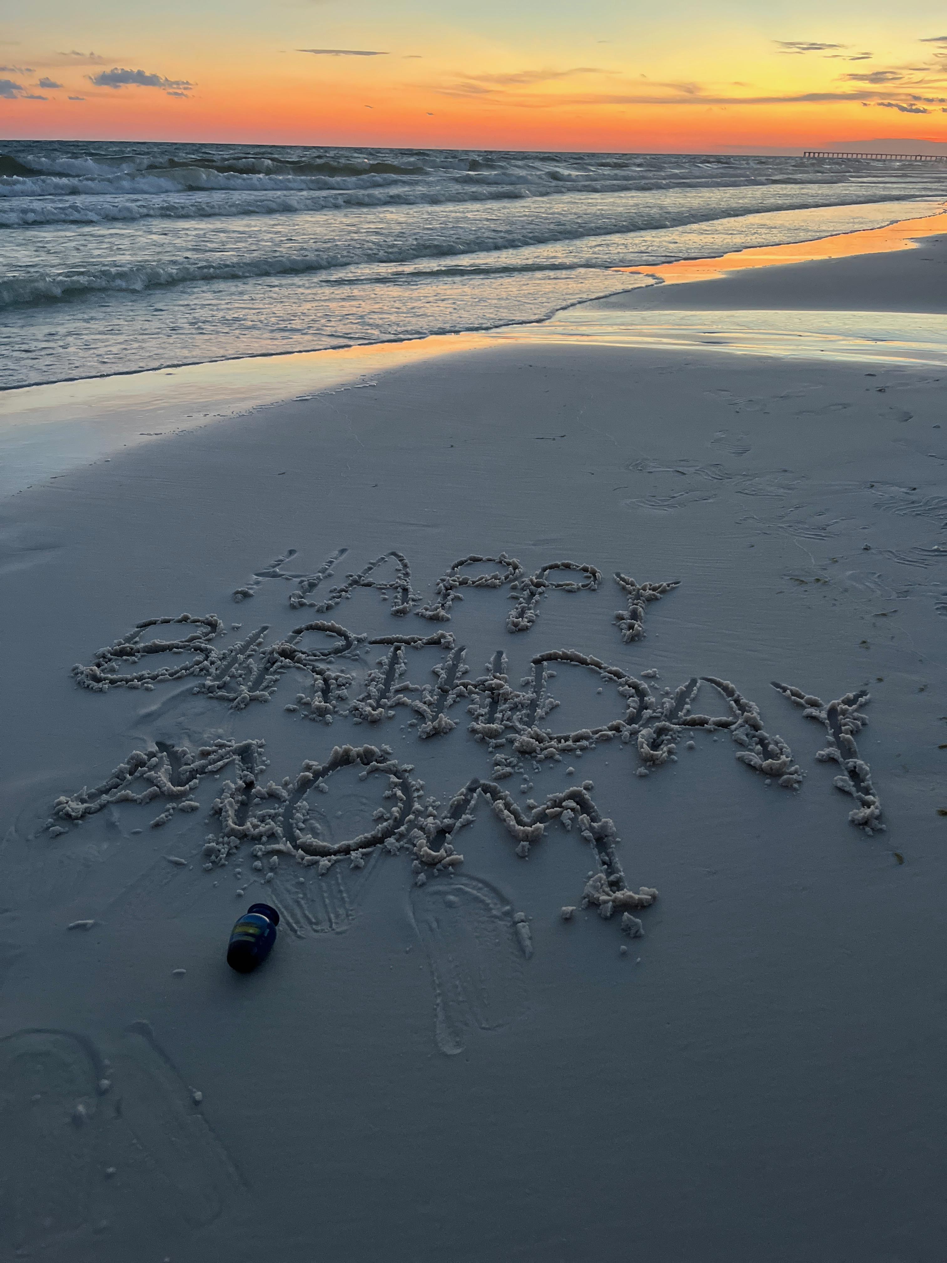 A birthday message is beautifully crafted in sand on the beach as waves crash gently.