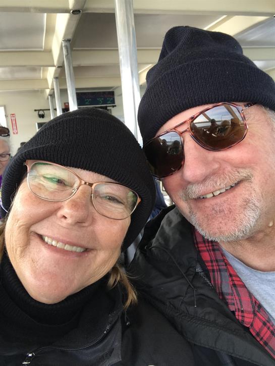A happy couple smiles while wearing winter hats and sunglasses on a ferry ride.