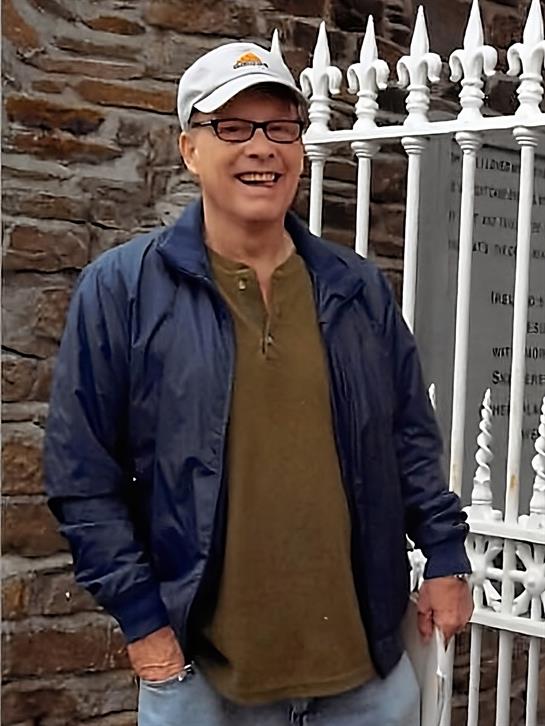 A man stands smiling by a white wrought iron fence, enjoying a warm day in a historic setting.