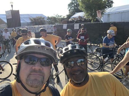 Two friends pose for a selfie while others get ready for a cycling event outdoors.