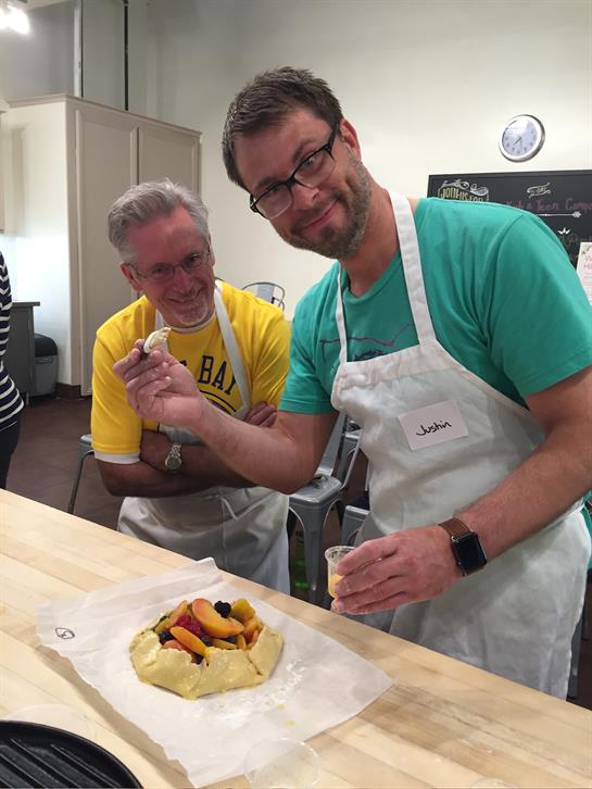 Two men enjoy making a fruit galette during a cooking class in a bright kitchen.
