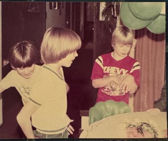 Three children engage in playful activity around a table filled with food during a family gathering.