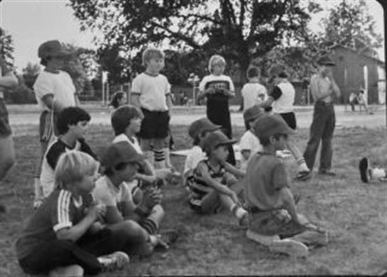 Children sit on the grass, watching a coach give instructions at a sports event.