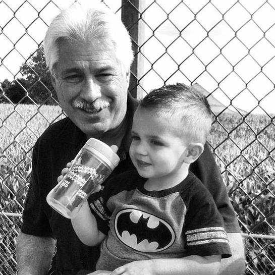 Elderly man sits with a young boy, both smiling and enjoying a refreshing drink outdoors.