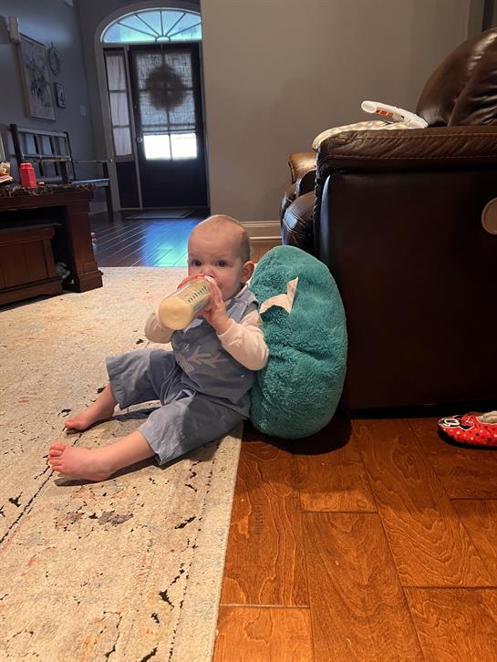 A baby sits comfortably on the floor, sipping from a bottle while leaning against a cushion.