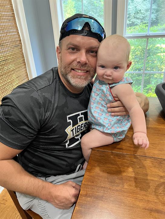 A father sits with his baby on his lap in a cozy home setting, both smiling happily.