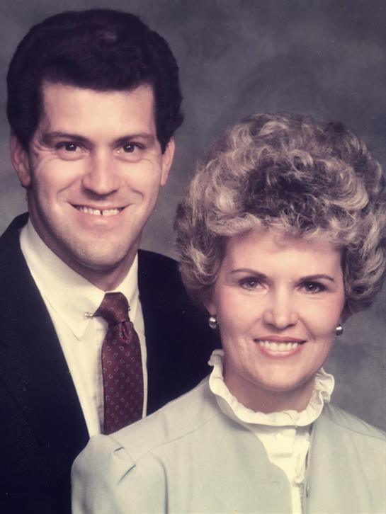 A couple stands close together, both smiling in formal attire, capturing a moment from the 1980s.