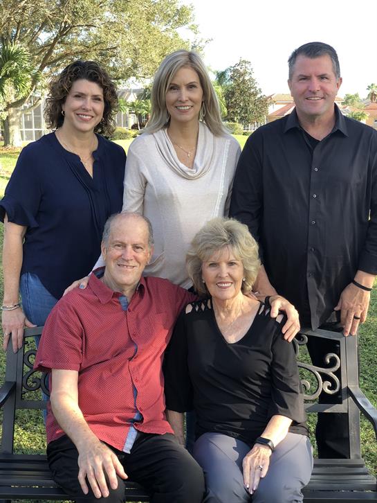 Five adults smile while sitting and standing together outdoors in a park setting.