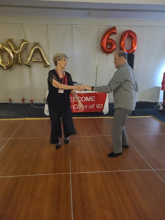 A couple dances joyfully at a reunion, surrounded by festive decorations and a welcoming banner.
