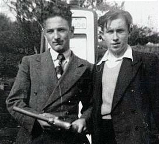 Two men stand together, one holding an object, with a gas pump behind them.