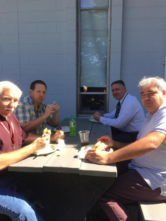 Four men share a meal at a picnic table while enjoying the warm sun and each other's company.