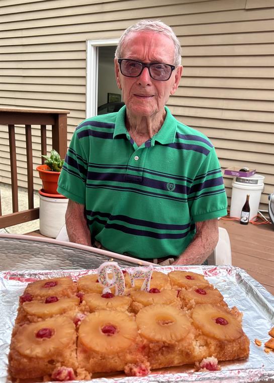 An elderly man smiles at a table with a cherry-topped pineapple upside-down cake.