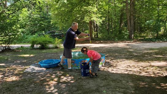 A father assists his young son in playing with water in a backyard setting on a sunny day.