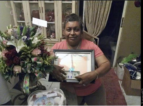 A woman stands in her living room showcasing a framed certificate, surrounded by flowers.