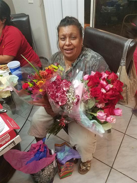 A smiling woman holds bright flower bouquets, surrounded by family at a special gathering.