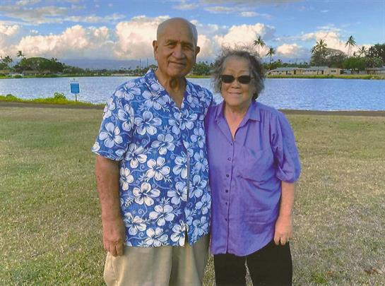 An elderly couple stands happily beside each other with a scenic lake backdrop and clouds.