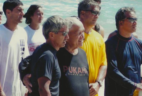 Two elderly men pose closely together while a group stands on the beach, enjoying a sunny day.