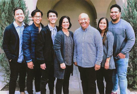 A cheerful family gathers outdoors, smiling and dressed casually in front of a stone archway.