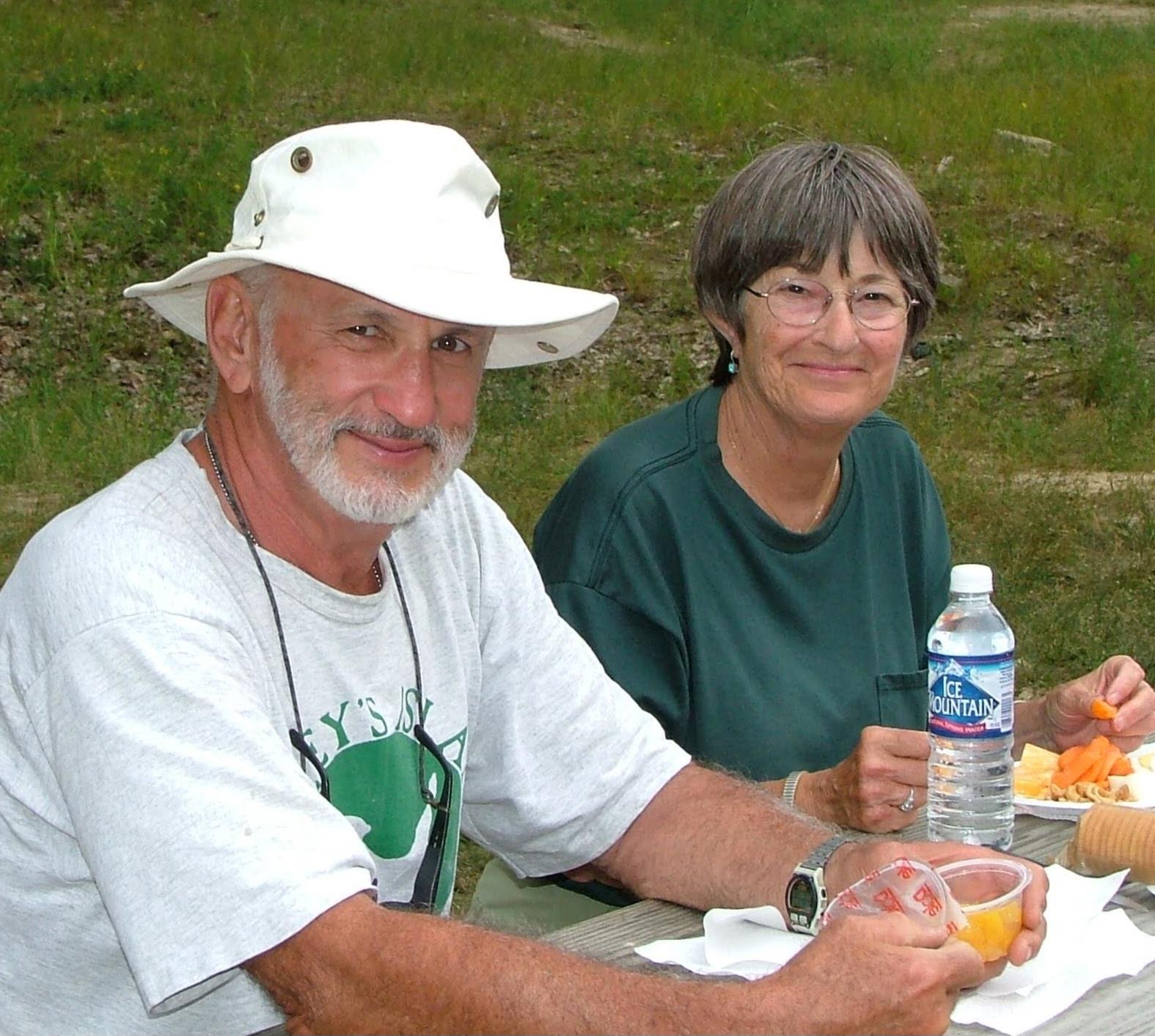 A couple sits at a picnic table, sharing food and drinks while enjoying nature together.