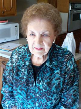 A senior woman stands in a cozy kitchen, smiling warmly while wearing a colorful blouse.