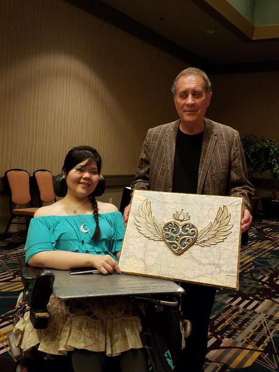 A woman in a wheelchair enjoys her time as she holds a heart design with an artist at a gathering.