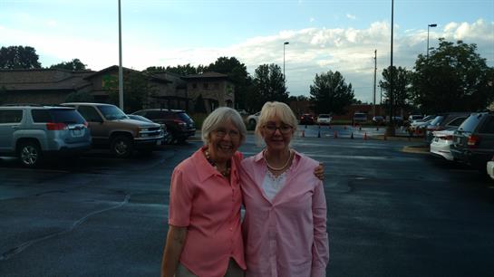 Two women stand together smiling in a parking lot under a bright afternoon sky.