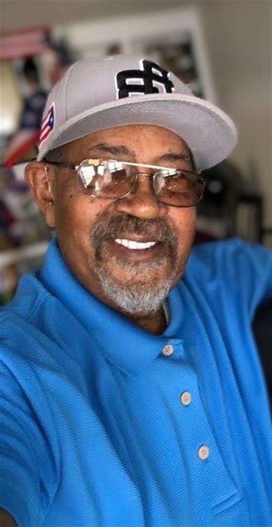 An older man in glasses and a cap smiles warmly in a cozy indoor selfie.