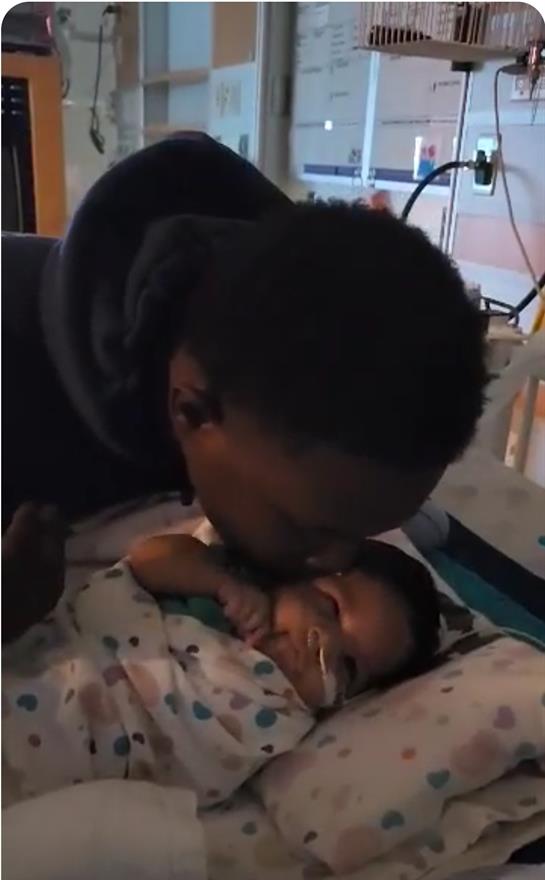 A father gently kisses his newborn child lying in a hospital bed, creating a tender moment.