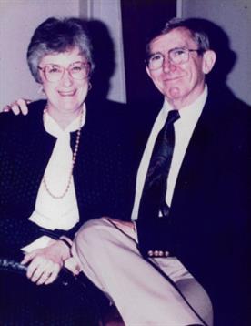 Elderly couple smiles warmly in formal attire, sharing an affectionate embrace.