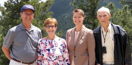 Four friends pose together outdoors, surrounded by trees and mountains in bright sunshine.