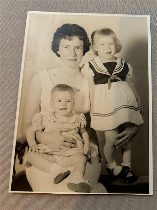 A smiling mother holds one daughter in her lap while standing next to another.