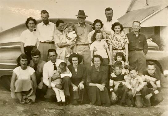 A large family gathers outdoors, smiling and posing near a vintage car on a sunny day.