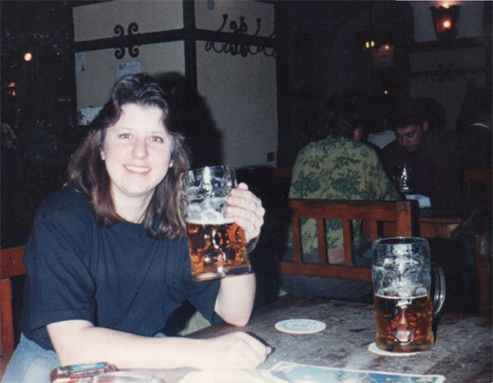 A woman smiles while holding a large beer in a lively pub filled with friends.