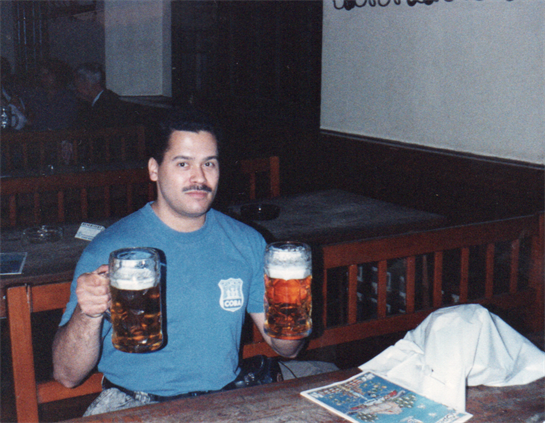 A man in a blue shirt holds two large mugs of beer at a wooden pub table, smiling.