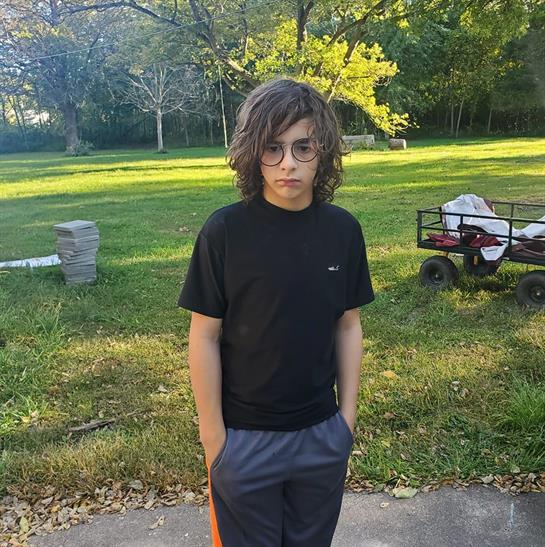 A boy with curly hair and glasses stands in a green park, surrounded by nature on a sunny day.
