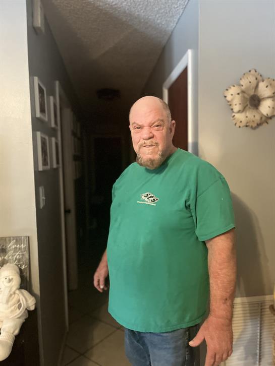 A man in a green shirt stands in a hallway, expressing a strong emotion while looking at the camera.