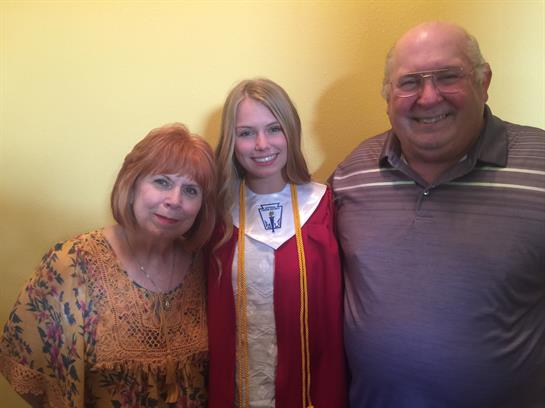 A student wearing a graduation gown poses with her smiling grandparents in a cozy setting.