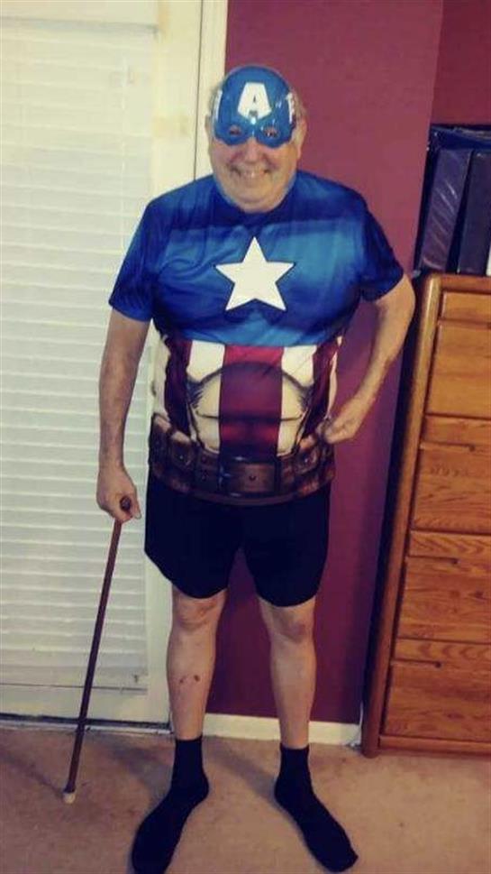 An older man dressed in a superhero outfit stands proudly in his home, smiling.
