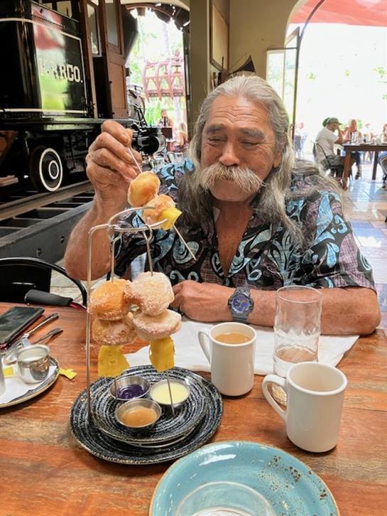 A man with long hair and a mustache savoring an elaborate dessert while seated indoors.