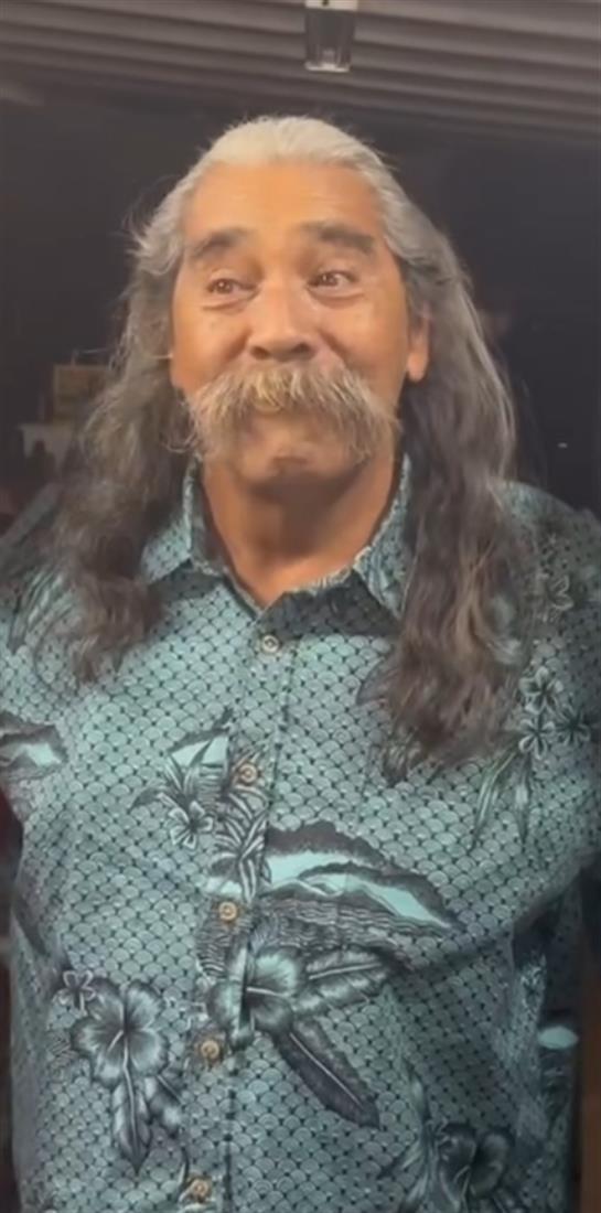 A man with long hair and a friendly smile wears a floral shirt while chatting in a warm environment.