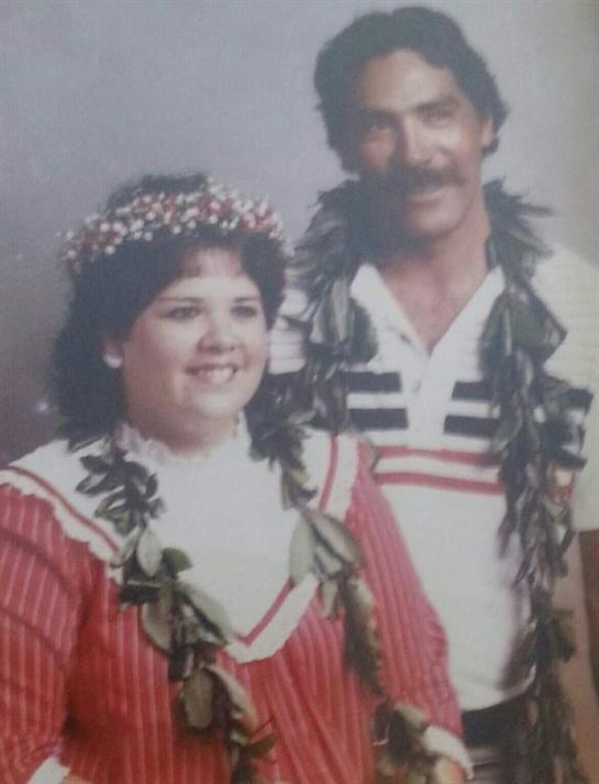 A couple dressed in vibrant traditional outfits, adorned with floral leis, smiles joyfully.