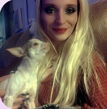 A woman with long blonde hair smiles while cuddling a small dog on a cozy couch.
