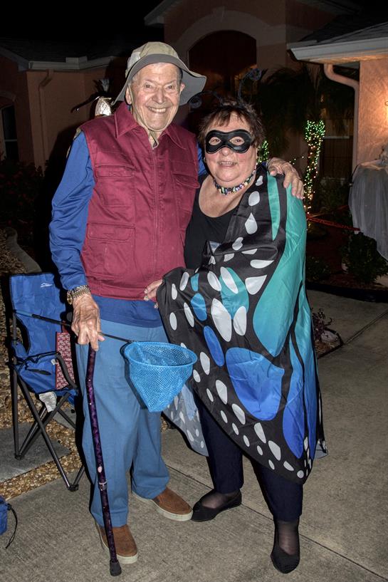 An elderly couple smiles in festive costumes at a neighborhood celebration.